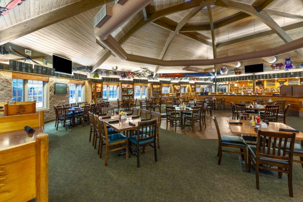 The image shows a spacious, rustic-style restaurant or bar with wooden furniture, tables set, and a visible bar area in the background.