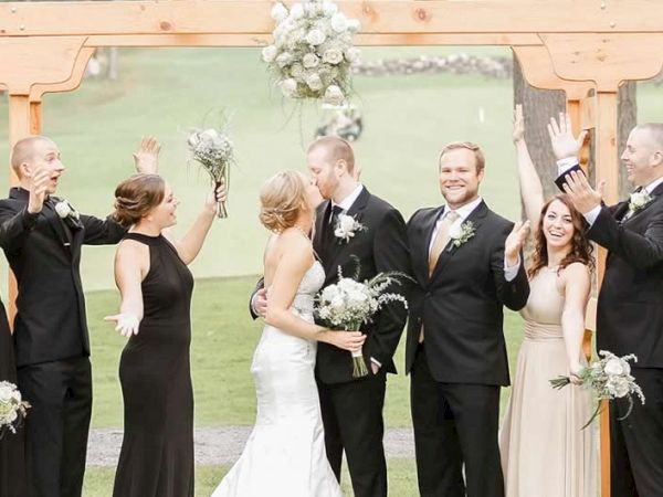 The image shows a bride and groom kissing under a wooden arch with a joyous wedding party surrounding them, raising their hands and celebrating.