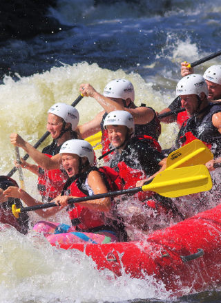 A group of people wearing life jackets and helmets are white-water rafting in a red inflatable raft, splashing through rapids and paddling together.