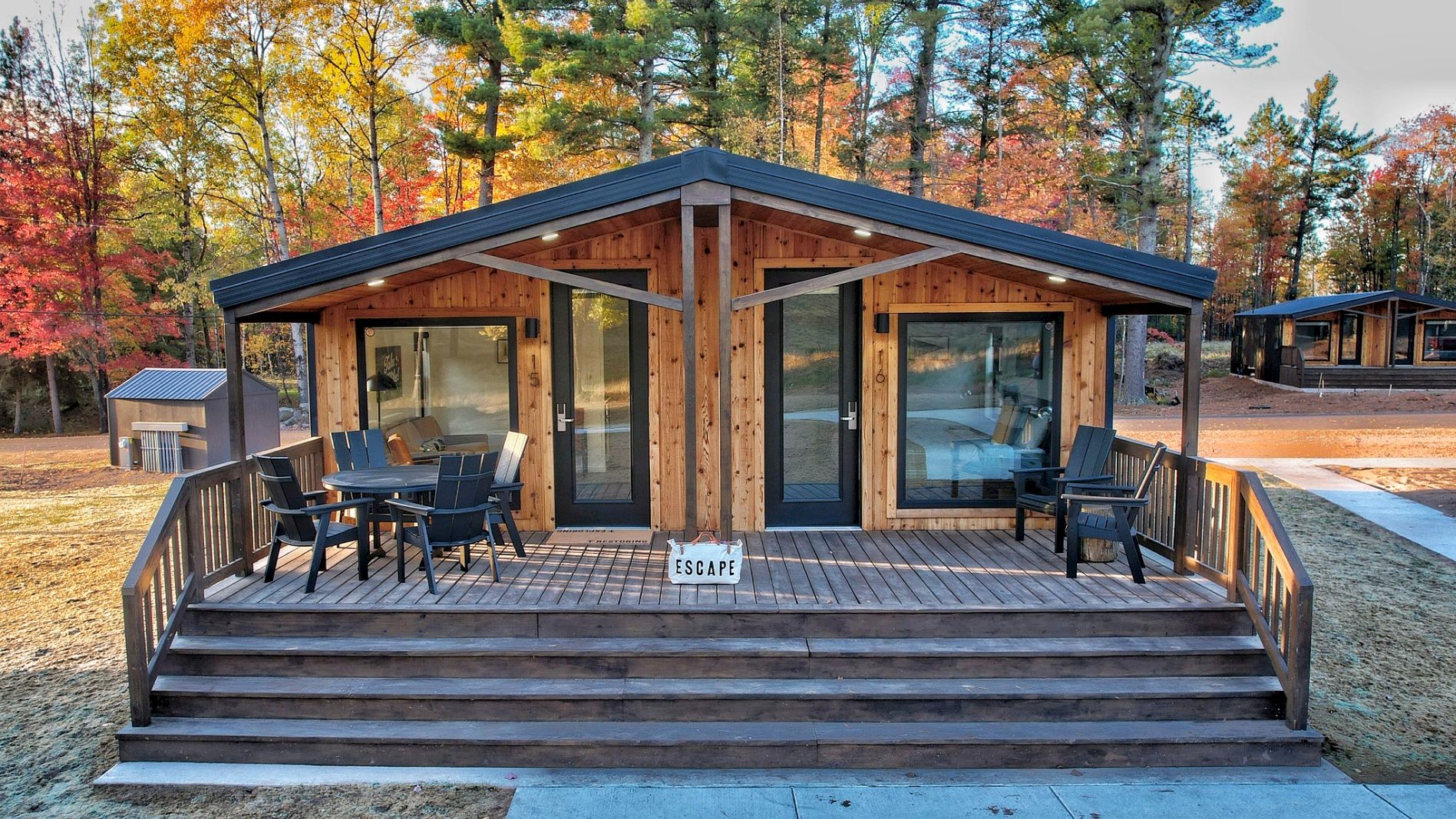 A cozy wooden cabin with a porch, two seating areas, surrounded by trees in autumn colors, offering a tranquil escape.
