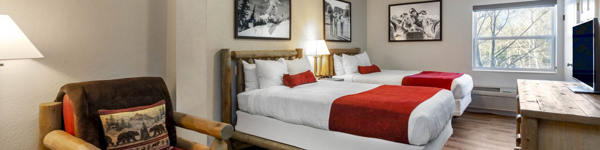 A cozy hotel room features two queen beds with red throws, a rustic armchair, a TV on a wooden dresser, wall art, and a window with natural light.