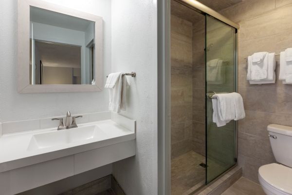 The image shows a modern bathroom with a sink, mirror, and shower. It features a glass shower door and a toilet with neatly folded towels.