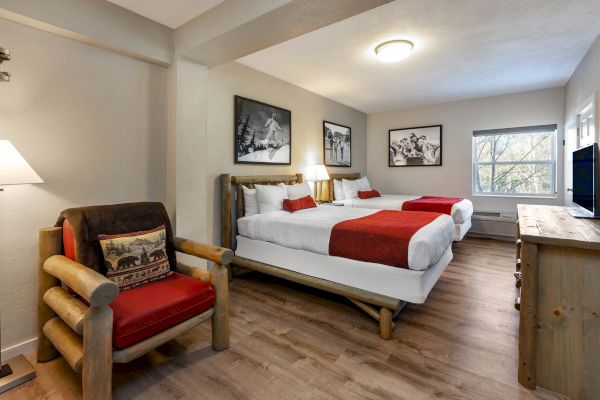 This image shows a cozy bedroom with two beds, red accents, a wooden armchair, framed pictures, a window, a lamp, and a TV on a dresser.