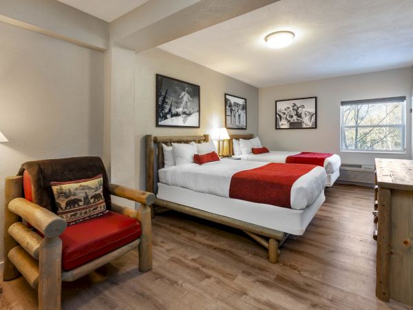 This image shows a cozy bedroom with two beds, red accents, a wooden armchair, framed pictures, a window, a lamp, and a TV on a dresser.