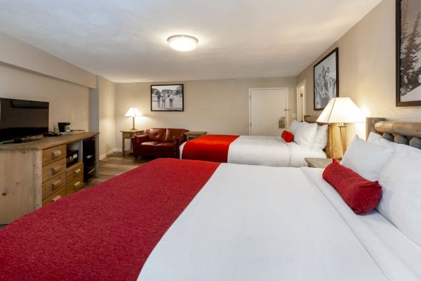 A hotel room with two large beds, red accents, a TV on a wooden dresser, a leather chair, and framed photos on the walls.