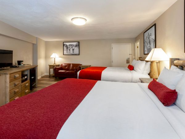 A hotel room with two large beds, red accents, a TV on a wooden dresser, a leather chair, and framed photos on the walls.