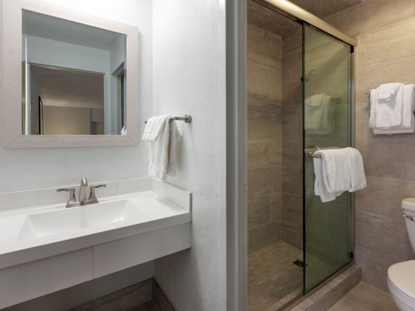 The image shows a modern bathroom with a sink and mirror, a towel rack with folded towels, a glass-enclosed shower, and a toilet.