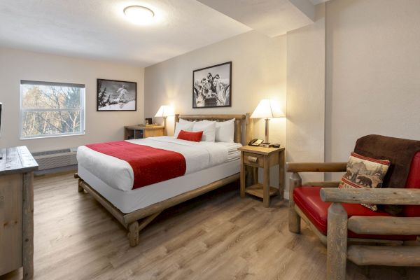 A cozy hotel room features a king-size bed, red accents, rustic wooden furniture, framed art, a cushioned chair, and a window with natural light.