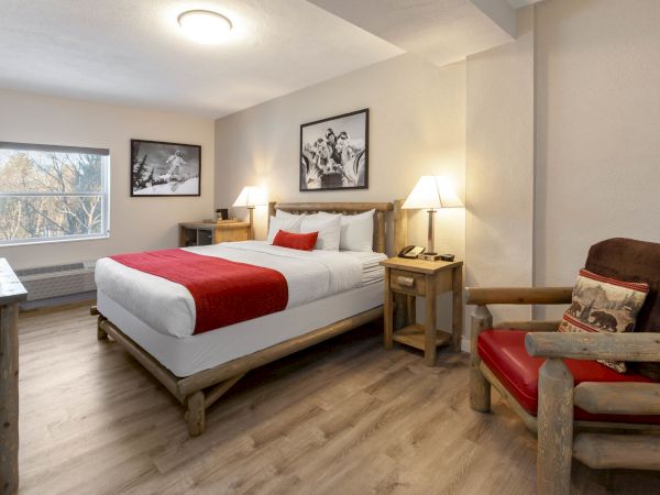 A cozy hotel room features a king-size bed, red accents, rustic wooden furniture, framed art, a cushioned chair, and a window with natural light.