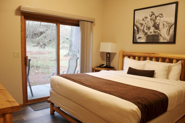 A cozy bedroom with a neatly made bed, a large framed picture above, a nightstand with a lamp, and a glass door leading outside.