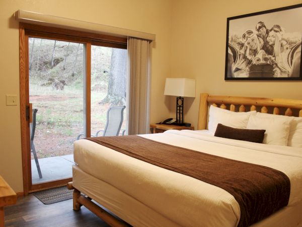 A cozy bedroom with a neatly made bed, a large framed picture above, a nightstand with a lamp, and a glass door leading outside.