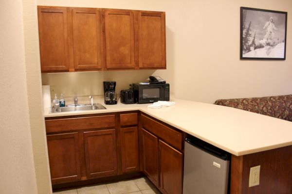 The image shows a small kitchen area with wooden cabinets, a sink, coffee maker, microwave, and mini-fridge.