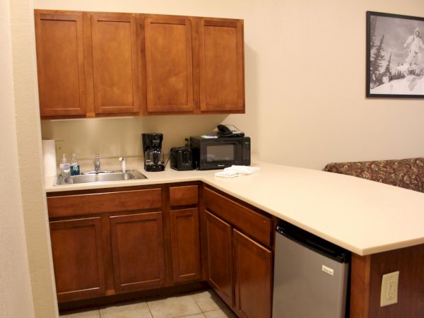 The image shows a small kitchen area with wooden cabinets, a sink, coffee maker, microwave, and mini-fridge.