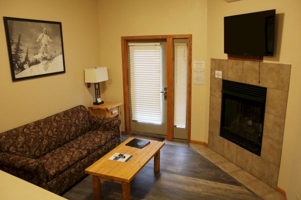 A cozy living room with a sofa, coffee table, lamp, wall art, television above a fireplace, and glass doors with blinds.