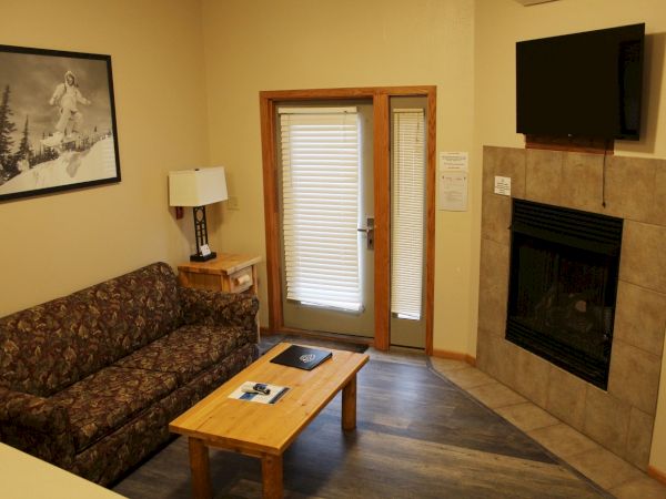 A cozy living room with a sofa, coffee table, lamp, wall art, television above a fireplace, and glass doors with blinds.