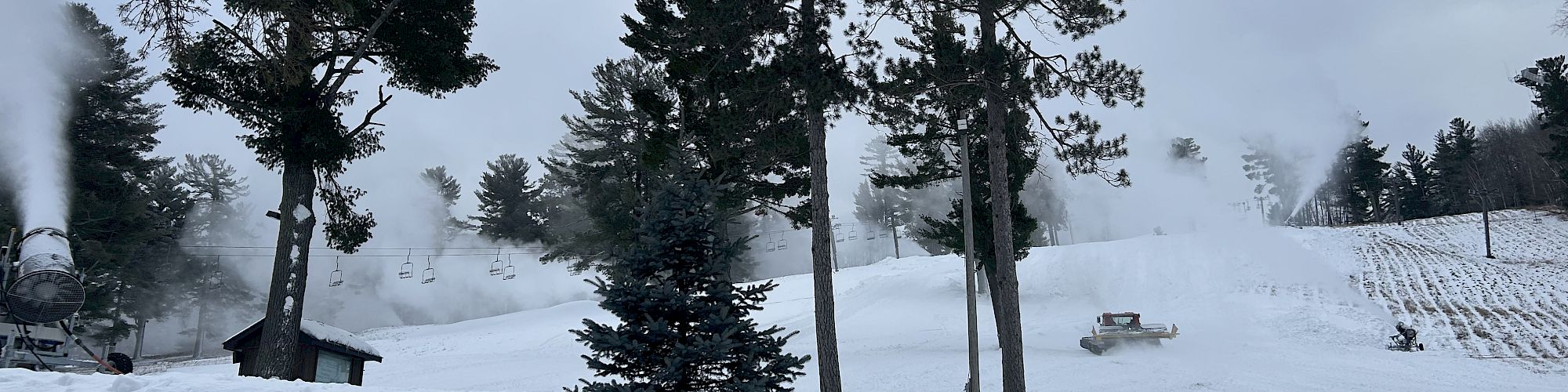 Snow-covered landscape with trees and snow machines creating artificial snow, under a cloudy sky.