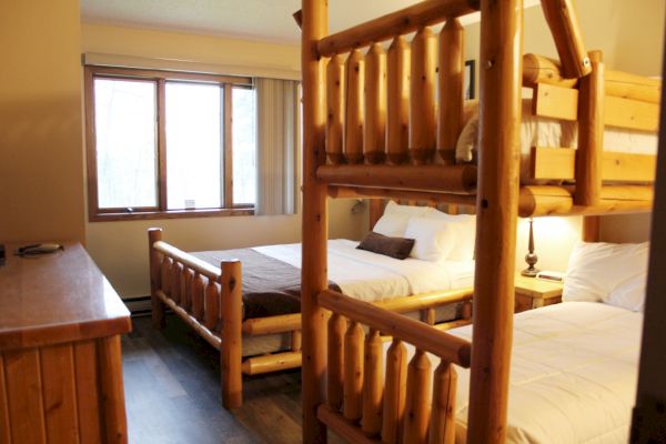 A cozy bedroom featuring wooden furniture with two single beds, a bunk bed, a window with blinds, and a wooden dresser in a rustic style.