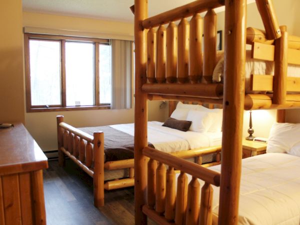 A cozy bedroom featuring wooden furniture with two single beds, a bunk bed, a window with blinds, and a wooden dresser in a rustic style.