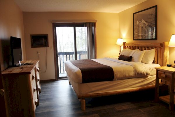 A cozy hotel room features a large bed, wooden furniture, a TV, lamps, and a sliding door leading to a balcony with an outdoor view.