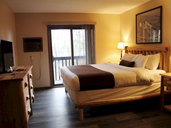 A cozy hotel room features a large bed, wooden furniture, a TV, lamps, and a sliding door leading to a balcony with an outdoor view.