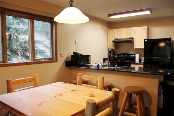 A cozy kitchen with wooden furniture, a dining table, and stools. It includes a stove, refrigerator, microwave, and a window with an outside view.