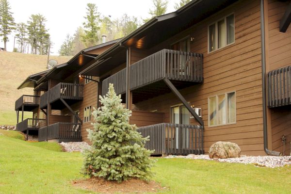 A row of brown wooden buildings with balconies and multiple windows, surrounded by green grass, trees, and rocks, under a clear sky.