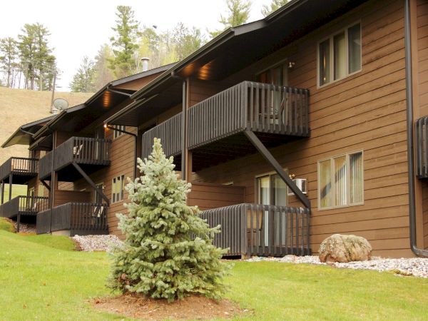 A row of brown wooden buildings with balconies and multiple windows, surrounded by green grass, trees, and rocks, under a clear sky.