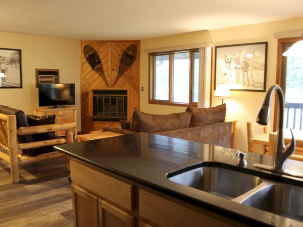 A cozy living room with rustic decor, featuring a kitchen counter, wooden furniture, fireplace, wall art, and a TV, all bathed in natural light.