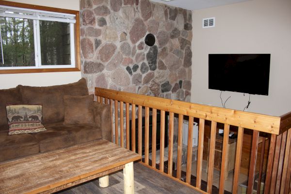 A cozy living area with a brown sofa, rustic coffee table, stone accent wall, mounted TV, wooden handrail, and window showing outdoor greenery.