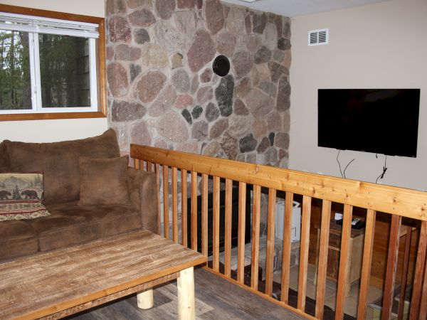 A cozy living area with a brown sofa, rustic coffee table, stone accent wall, mounted TV, wooden handrail, and window showing outdoor greenery.