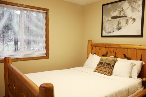 A cozy bedroom with a wooden bed frame, white bedding, a decorative pillow, a window, and a wall-mounted picture of a skier.