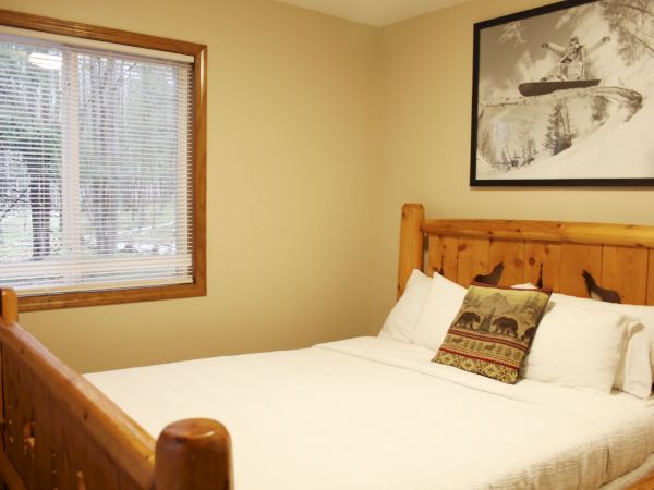 A cozy bedroom with a wooden bed frame, white bedding, a decorative pillow, a window, and a wall-mounted picture of a skier.