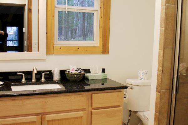 The image shows a bathroom with a wooden-framed window, a vanity with a sink and mirror, a toilet, and a glass-enclosed shower.