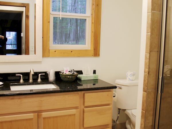 The image shows a bathroom with a wooden-framed window, a vanity with a sink and mirror, a toilet, and a glass-enclosed shower.