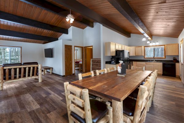 A cozy open-concept cabin interior with wooden beams, a large dining table, kitchen appliances, comfortable seating, and wood accents throughout.