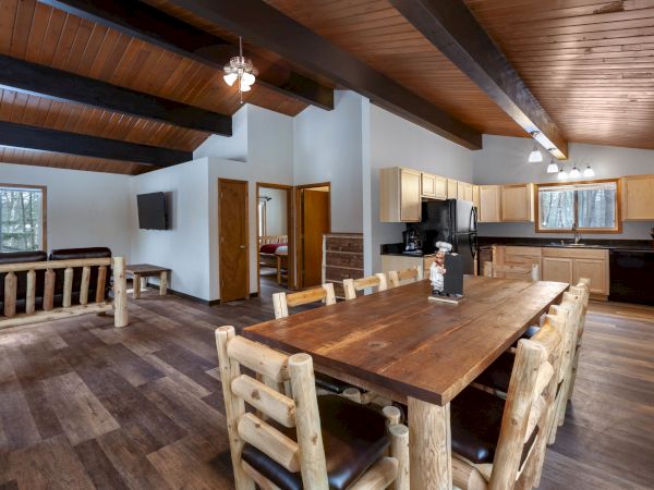 A cozy open-concept cabin interior with wooden beams, a large dining table, kitchen appliances, comfortable seating, and wood accents throughout.