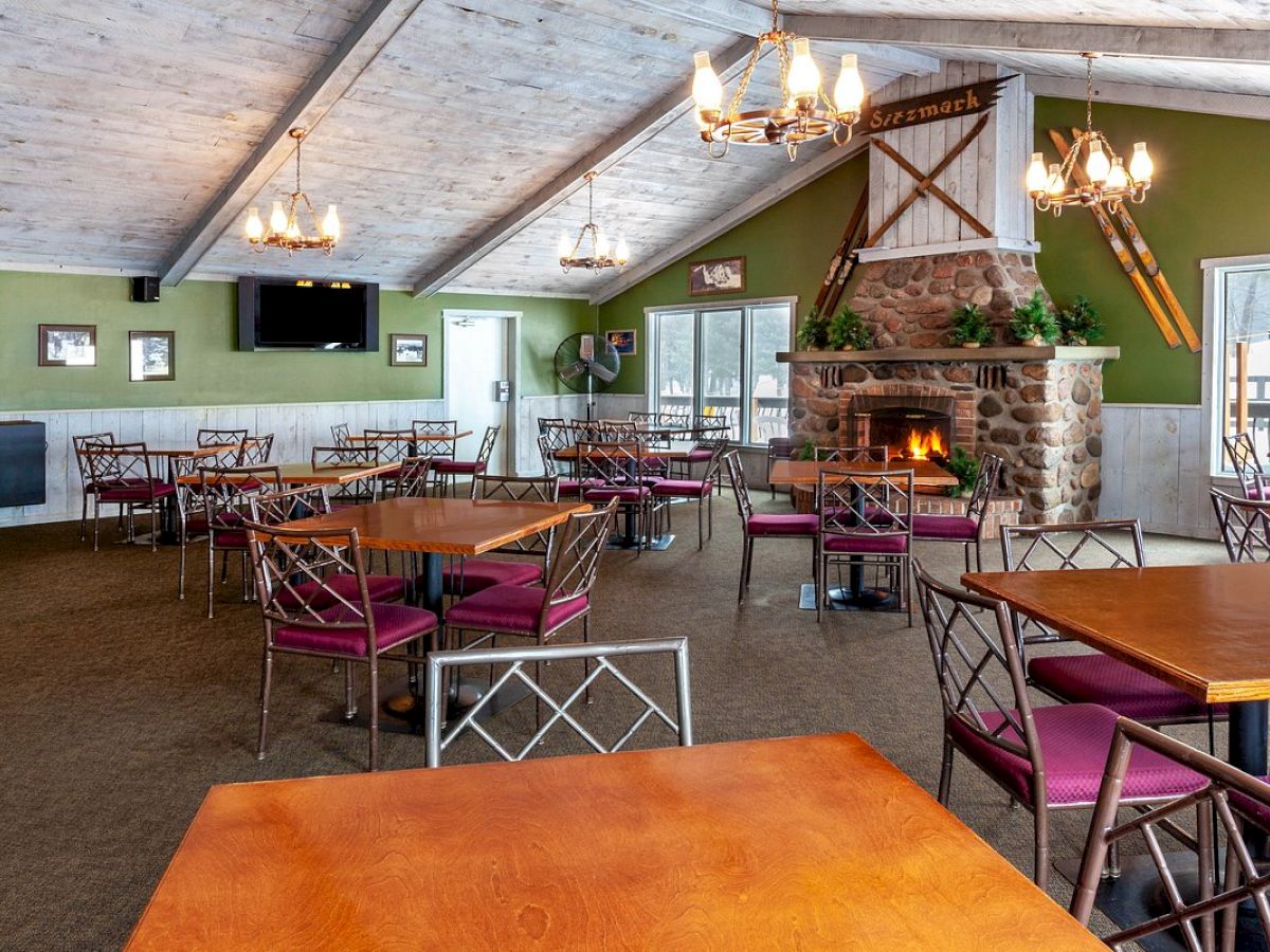 A cozy, empty restaurant with wooden tables, purple-cushioned chairs, chandeliers, a stone fireplace, and large windows is depicted in the image.