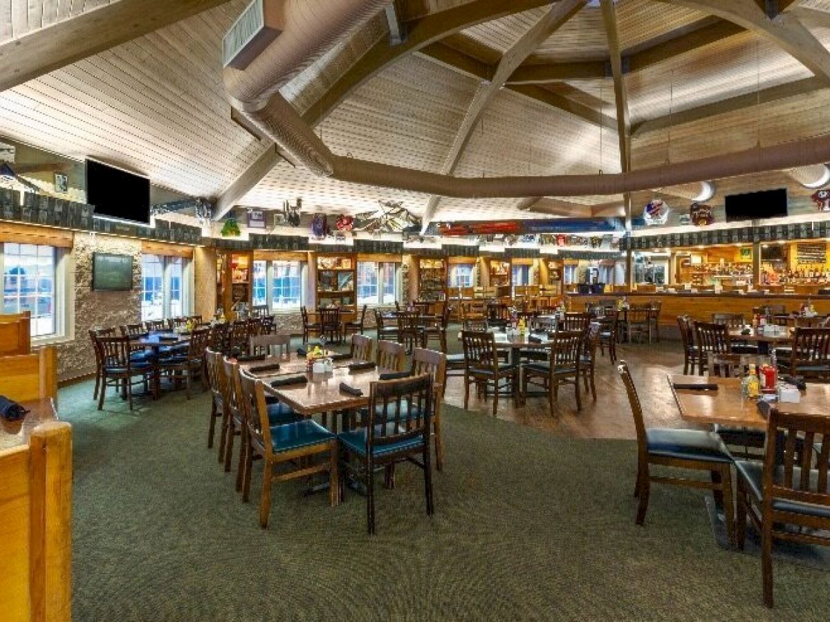 A spacious restaurant interior with wooden tables and chairs, a bar area in the background, and a vaulted ceiling with skylights, all well-lit and cozy.