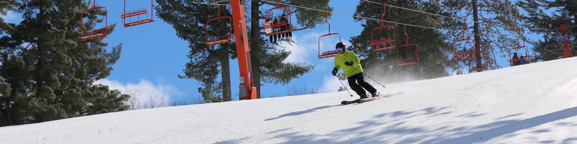 A skier in a bright green jacket descends a snowy slope surrounded by trees, with red chairlifts in the background against a clear blue sky.