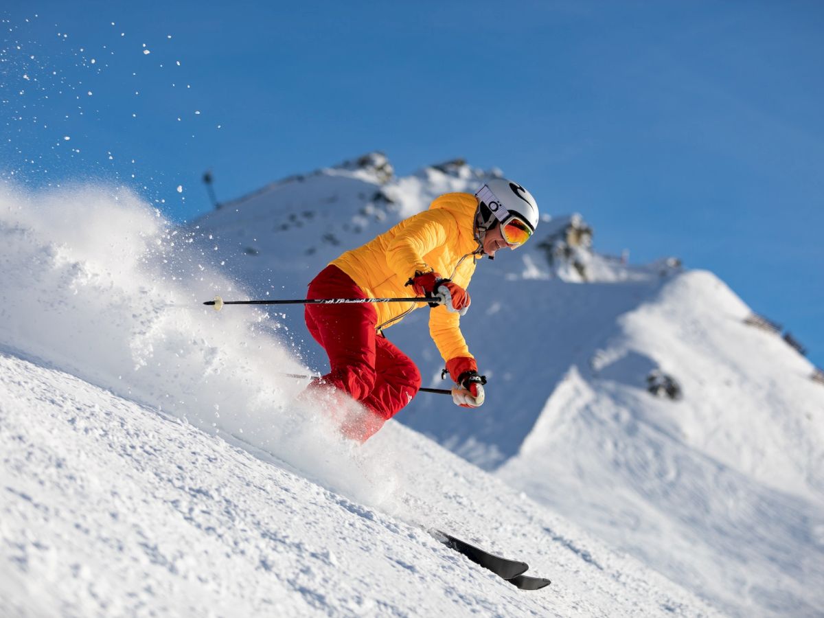 A skier wearing a yellow jacket and red pants is descending a snowy mountain slope, kicking up powder and gripping ski poles on a sunny day.