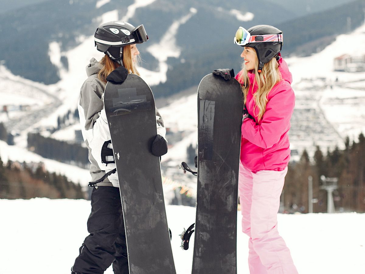 Two people in ski gear holding snowboards stand on a snowy mountain with ski trails and chairlifts in the background.