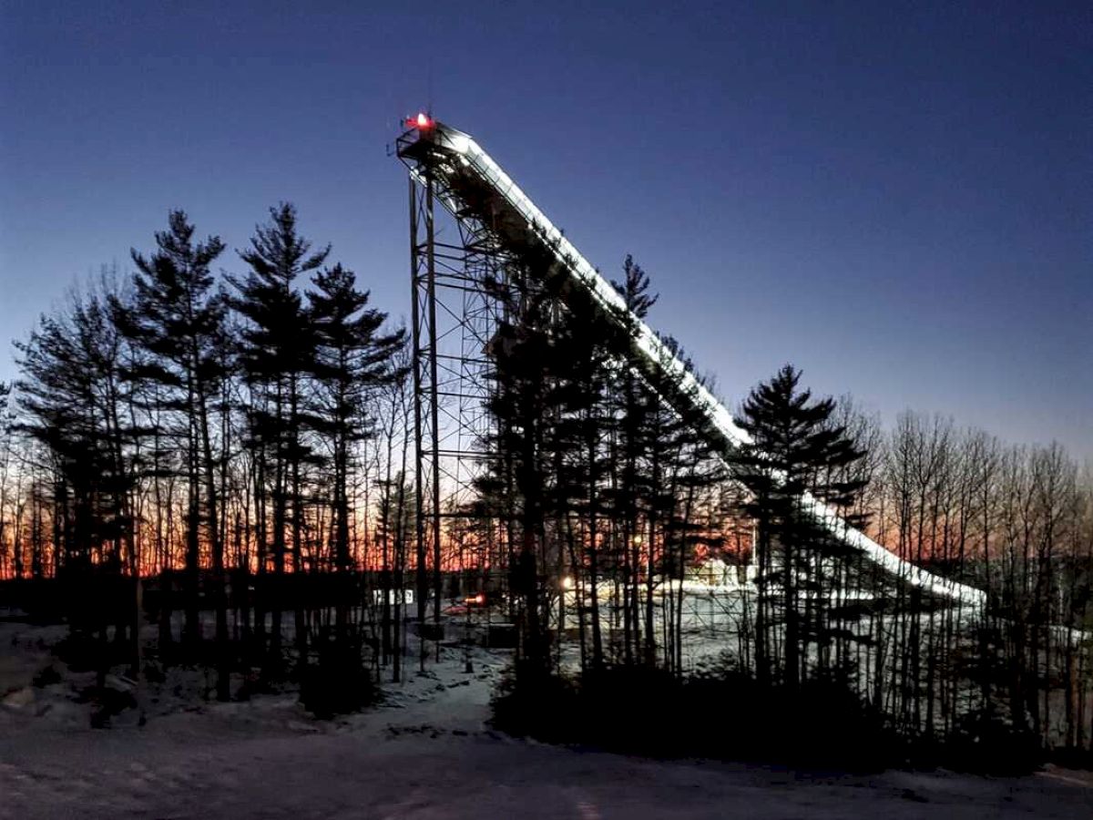 This image shows a snow-covered ski jump structure at dusk, surrounded by tall trees. The sky has a gradient of dark blue to orange.