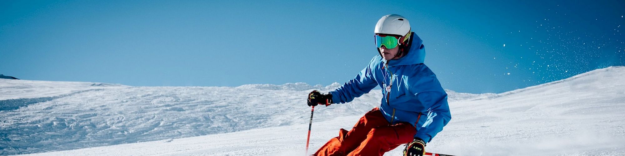 A person in a blue jacket and red pants is skiing on a snow-covered slope with clear blue skies in the background, wearing a helmet and goggles.