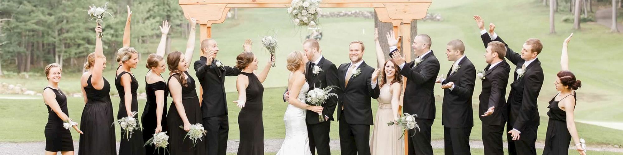 A wedding party celebrates under an arch in an outdoor setting, with bridesmaids and groomsmen raising their arms while the couple kisses.