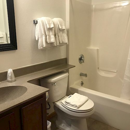 This image depicts a clean, modern bathroom with a sink, mirror, towels, toilet, and a white bathtub with a shower curtain.