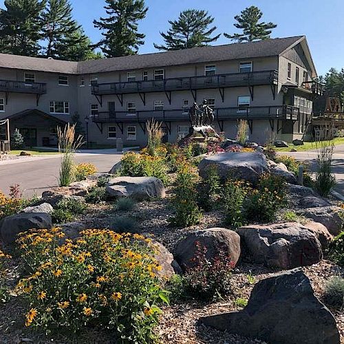 The image shows a landscaped garden with rocks and flowers in front of a multi-story building surrounded by tall trees.