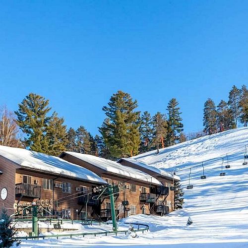 A snowy ski resort with buildings, ski lifts, and fir trees under a clear blue sky, showing a serene, well-maintained winter landscape.