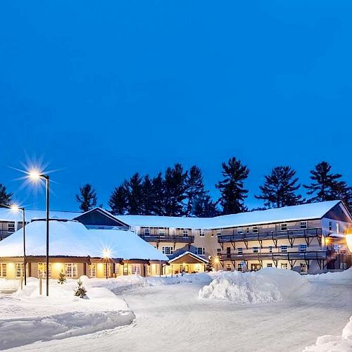 A snow-covered building at dusk, with lights illuminating the area, surrounded by trees under a clear blue sky ending the sentence.