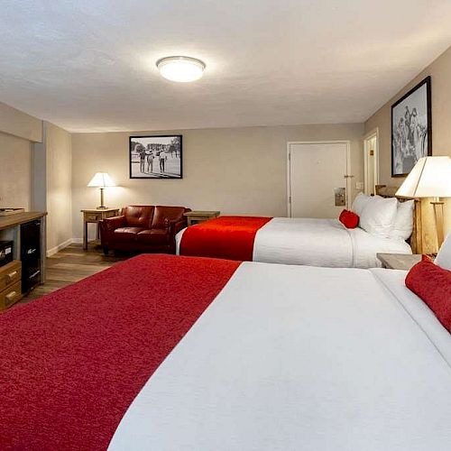 A hotel room with two double beds, red accents, a TV, dresser, armchair, side table with a lamp, and framed pictures on the walls.