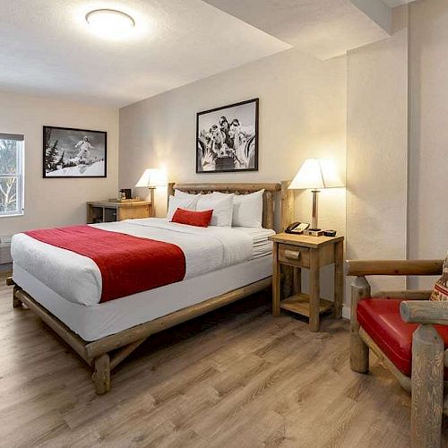 A cozy hotel room with a large bed, red accents, wooden furniture, framed pictures, a window with a view, a nightstand, and a red cushioned chair.